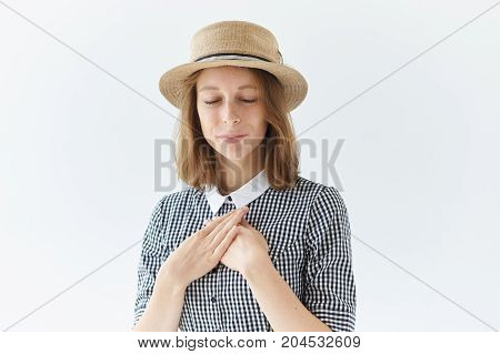 Grateful young woman in round hat holding both hands on chest and closing eyes expressing gratitude and appreciation. Friendly sincere joyful girl saying thanks her heart is full with love