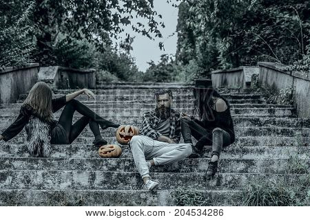 Halloween holiday celebration concept. Halloween people and jack o lanterns. Friends on stony stairs. Women with pumpkin heads. Bearded man sitting with girls.