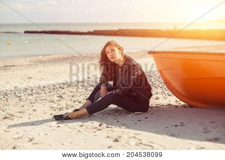 A girl sideways sitting near a red boat on the beach by the sea with beautiful sunset on the background. Woman in jeans pullover and jacket on the sand