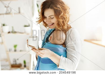 Joyful pretty mother with sleeping son in baby sling, holding cup of tea in hand and laughs chatting with her best friend on smartphone in light cozy room