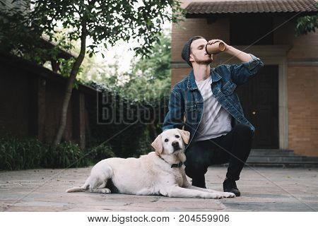 A young guy squatting near his best pet in the world and drinking tasty coffe from the brown cup while the golden labrador retreiver is sitting on the asphalt and looking to the camera.