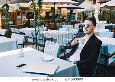 Nice picture of a promising young businessman sitting at the table in a big restaurant. He is drinking coffee and relaxing during the short break
