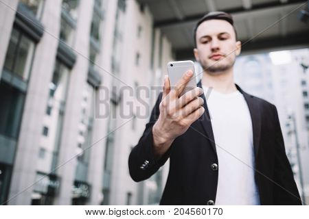 Attractive and young enterpreneur is standing near big office building and looking to his phone. There is an important information to be checked. This man looks serious but confident