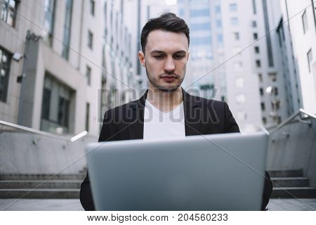 Tha portrait of a businessman sitting near big office building. He is working on the laptop very attentive and serious. Close up. Cur view