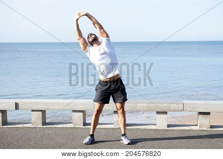 Serious young handsome strong man wearing sportswear, stretching body, doing side bend exercise and standing on bridge with sea in background. Front view.