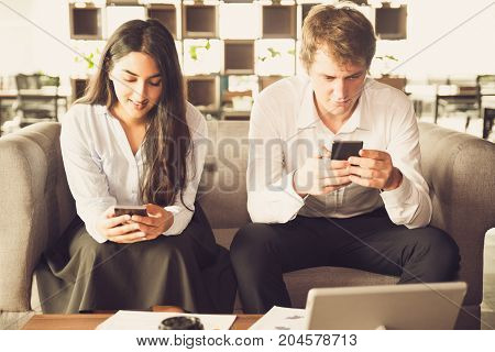 Addicted business people using gadgets in office. Concentrated young colleagues immersed in digital space. They reading message or surfing net while sitting on sofa. Technology concept