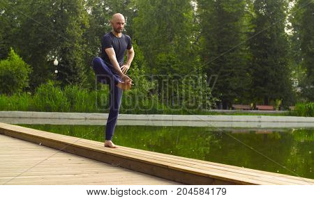 Strong man performing one-leg standing balance near the pond. Yoga asana - utthita hasta padagushthasana