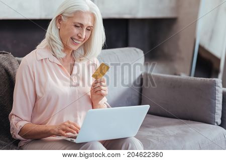 Purchase online. Waist up of a cheerful retired woman holding her credit card and using her laptop while enjoying online shopping