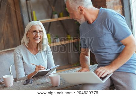 Healthy relationships. Cheerful delighted aged woman smiling and making notes while her pleasant husband using laptop