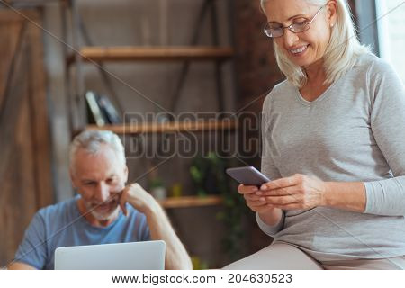 write me back. Cheerful beautiful aged woman using her smart phone and texting while her husband using the laptop in the background