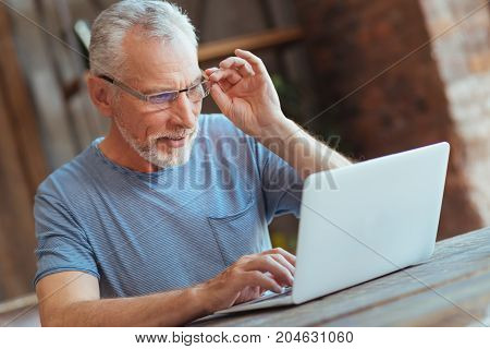Surf the Internet. Waist up of a cheerful aged man sitting at the table and using his laptop while touching his glasses