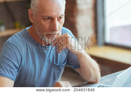 Let me think. Serious thoughful aged man sitting at the table and using laptop while mulling over his problems