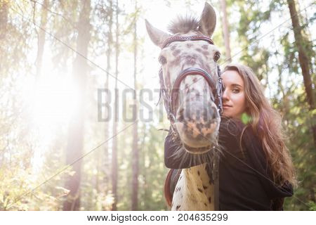 Young caucasian woman taking care of her horse, hugging his neck. Horse is white in brown spot. They arre walking in forest. Film effect
