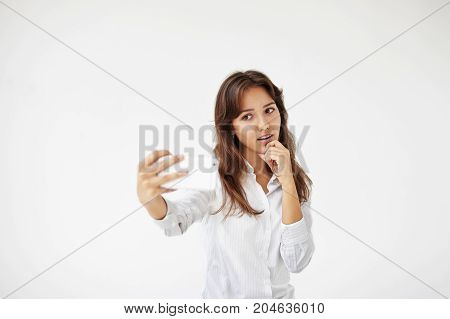 Collecting memories. Headshot of charming young woman employee in formal wear taking self pictures using smart phone. Beautiful Latin female worker posing at white office wall taking selfie