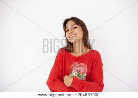 Waist up shot of happy cheerful young mixed race female wering red top holding tight big glass jar of colorful marmalades or dragee looking and smiling broadly at camera. Food sweets and sugar