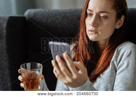 Unhealthy lifestyle. Cheerless moody upset woman using her smartphone and holding a glass with whisky while being alcohol addicted