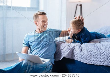 Fatherly affection. Pleasant happy father sitting on the bedroom floor with a laptop in his lap and ruffling the hair of his little son lying on the bed