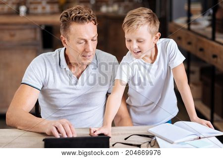 Helpful advice. Charming pre-teen boy giving his father a piece of advice regarding a tablet and pressing necessary icon on tablet while the father listening to him attentively