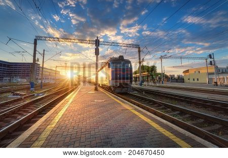 High speed passenger train on railroad track at sunset in Europe. Railway station with modern commuter train departure, blue sky with clouds and sun. Colorful Industrial landscape. Railway platform