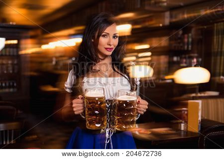 Beautiful female waitress wearing traditional dirndl and holding huge beers in a pub. Sexy brunette carries two glasses of beer between the tables in the bar
