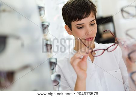 Pretty young woman is choosing new glasses at optics store to correct eyesight