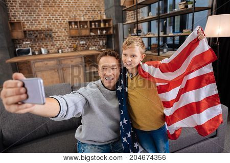 Happy state holiday. Cheerful young father sitting on the sofa and taking a selfie with his cute pre-teen son wearing an American flag, celebrating a state holiday