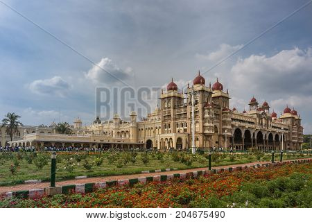 Mysore India - October 27 2013: Wide shot out of garden of South and East facades of Mysore Palace under cloudscape. Yellow building with towers and maroon domes. Flowers up front.