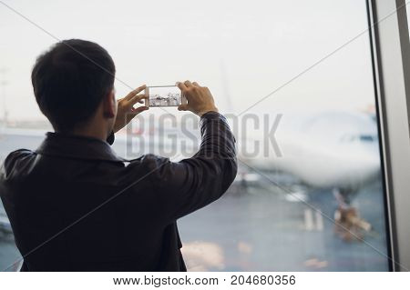 Handsome man standing beside glass wall in modern airport terminal, taking photo picture of airplane aircraft, travelling to other countires