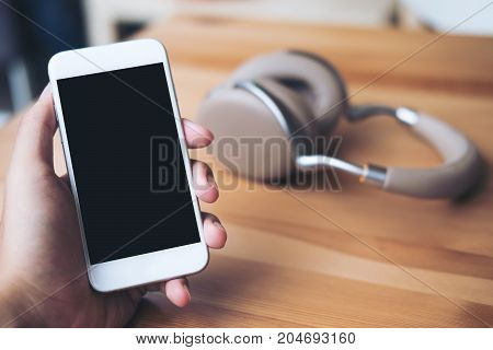 Mockup image of a man's hand holding white mobile phone with blank black screen and brown headphone on vintage wooden table in cafe