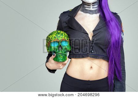 girl with makeup and purple hair holds a painted skull in her hand on a gray background. Mexican celebration of the Day of the Dead Dia de los Muertos and the Roman Catholic holiday All Souls Day