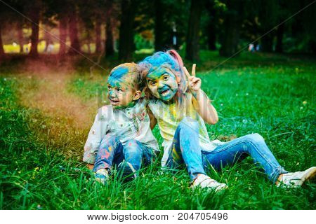 little girl with boy sit on the ground all dirty in holi paints in city park