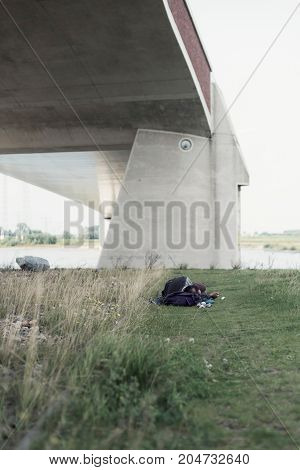 Rambler Sleeps In Grass Under Modern Bridge Near River.
