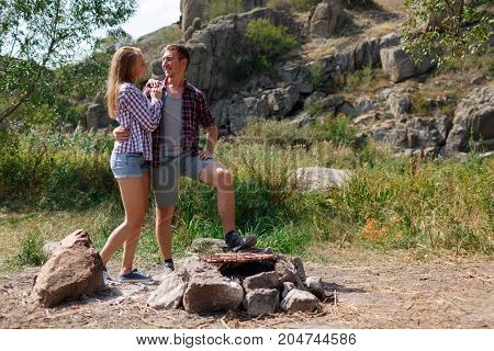 A young couple is traveling in the mountains. Summer journey of lovers, a guy frying meat at a bonfire for a girl. Healthy way of life, travel, summer vacation. The best pastime in the summer.