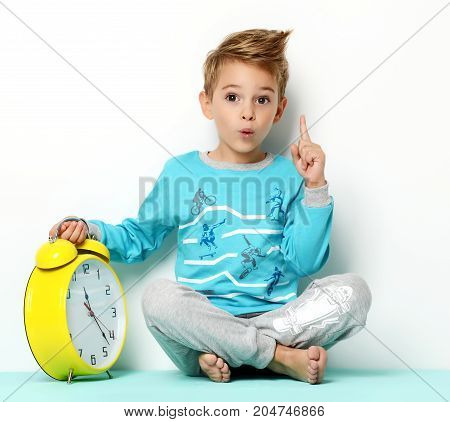 Happy young boy sitting in blue sweater thinking and looking up with big yellow clock pointing finger up on white background