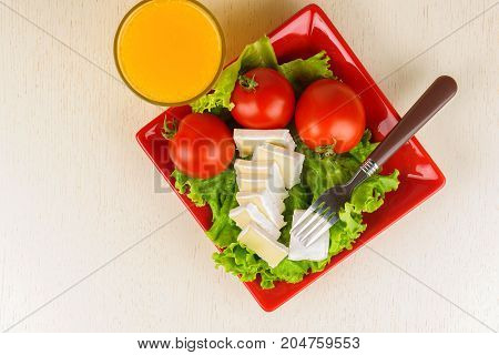 Tomatoes And Cheese Slices Lie On A Sheet Of Fresh Salad In A Red Plate. A Glass Of Orange Juice Sta