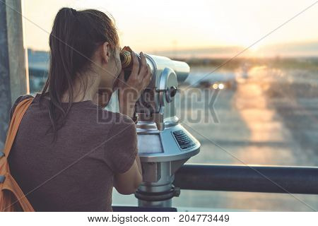 Girl at the airport window looking outside through a telescope