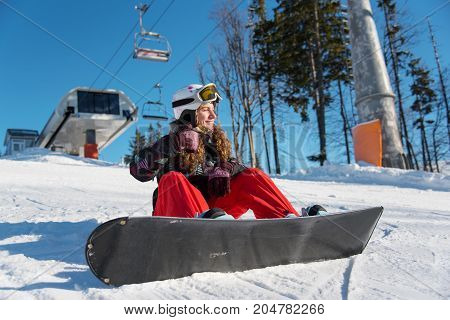 Smiling girl with snowboard sitting on the snow under a ski lift at ski resort and showing thumbs up gestures of good class
