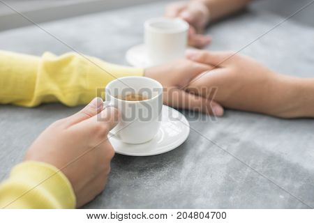 Close-up young couple is holding hands in the outdoor at coffee shop by intense focus on edge of coffee cup for make behind blurred.