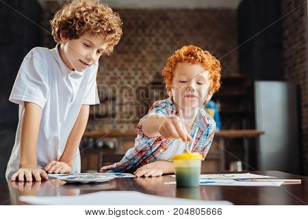 Next great artist. Cute redhead boy washing his painting brush while working on a new masterpiece while his older brother standing next to him and looking at his painting.