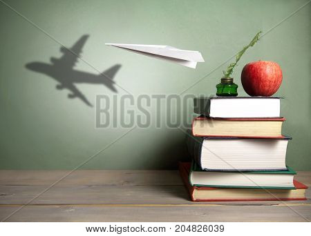 Paper plane with shadow of an aircraft next to a stack of books quill and apple
