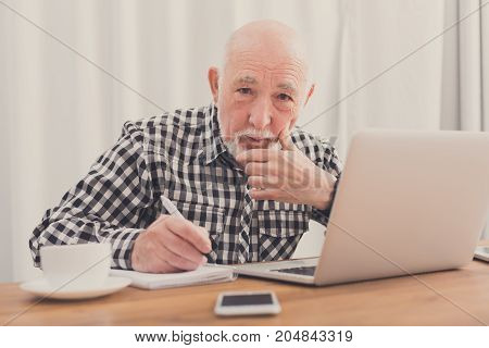 Mature man using laptop and writing in notepad at home desk. Education, recipe concept
