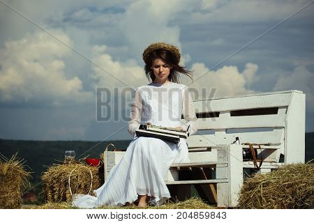 Woman typing on typewriter on cloudy sky. Girl in wreath and white dress on bench. Assistant or secretary working on nature on sunny day. Vintage equipment and archaism. Summer vacation concept.