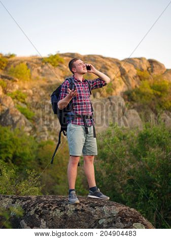A traveling young man, walking near the mountains and calling a phone on a light natural background. Sad, angry tourist talking on a phone with a bad connection. Camping problems concept. Copy space.