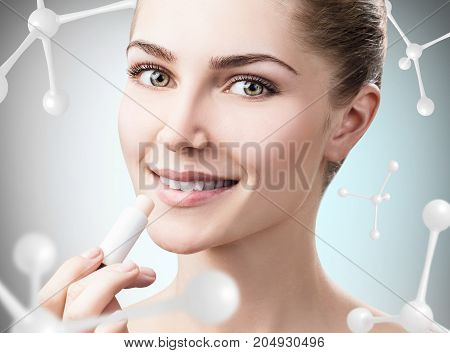 Beautiful young woman putting lipbalm among molecules over gray background. Innovation cosmetics concept.