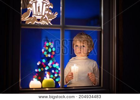 Beautiful little kid boy, standing by window at Christmas time and holding candle. Child waitng for Santa. With colorful lights from Christmas tree on background.