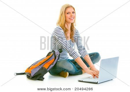 Teengirl sorridente seduto sul pavimento con zainetto e l'utilizzo di laptop isolata on white