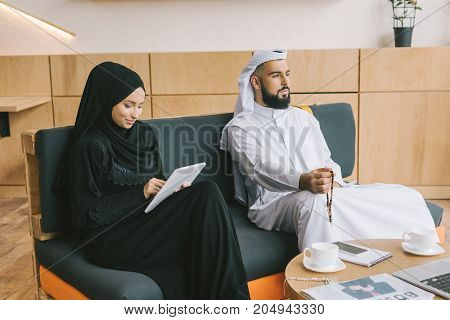 musilim woman sitting on couch and using tablet while her husband looking away thougtfully