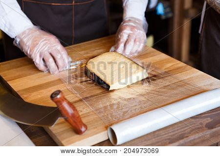Shop assistant wrapping a piece of cheese. Packing order at grocery shop in cling film on wooden table, background