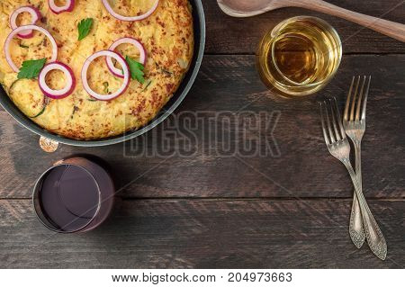 An overhead photo of a Spanish tortilla with potatoes and zucchinis in a tortillera, with glasses of red and white wine, and a place for text