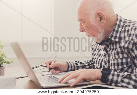 Mature man using laptop and writing in notepad at home desk. Education, recipe concept